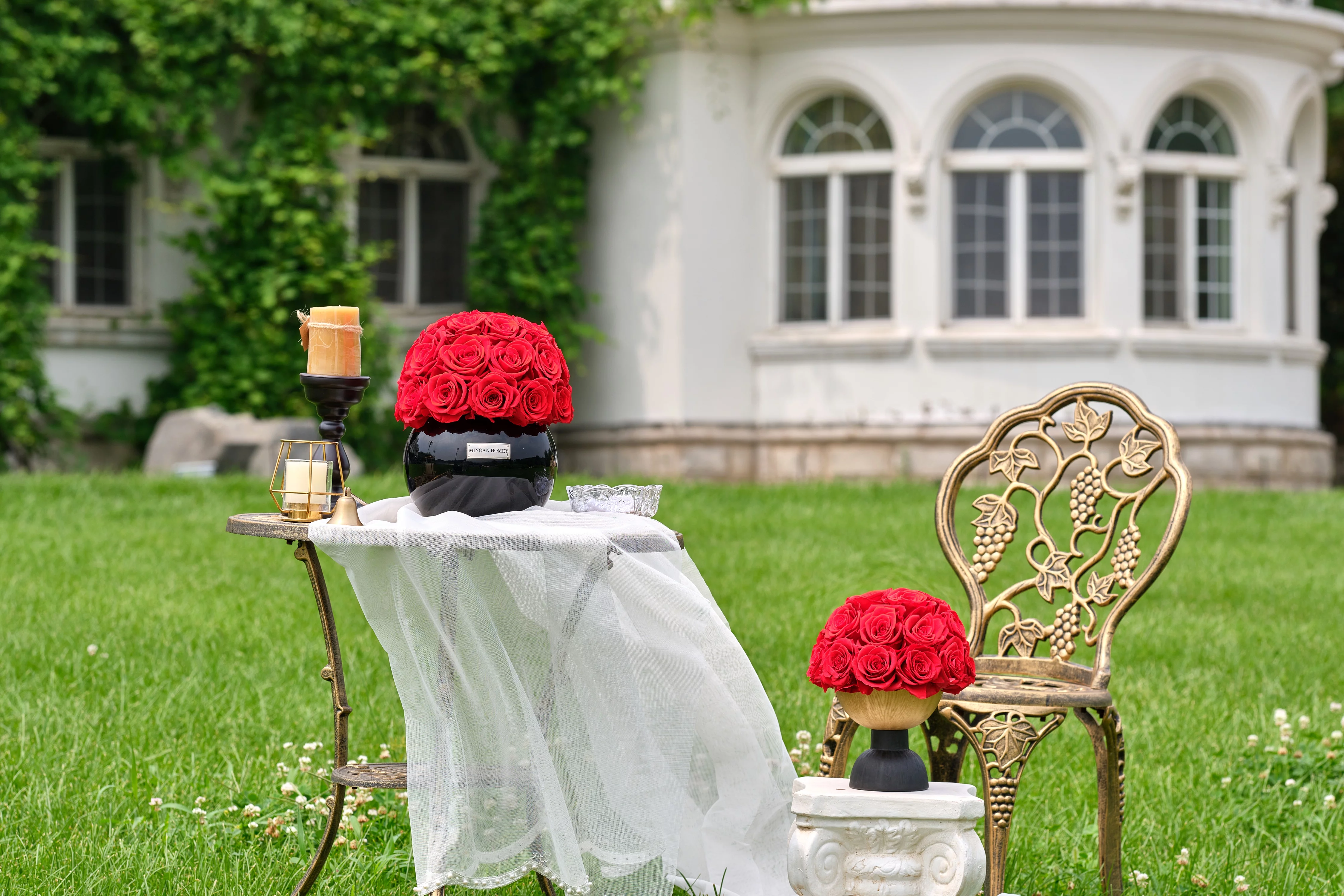 Elegant outdoor setting with white wrought iron chair and table. Black vases with red roses and lit candle create a floral-themed sophisticated display.